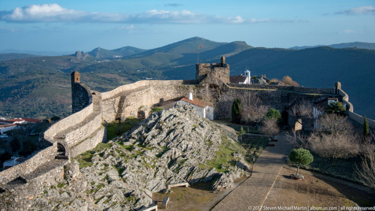 Elevated overview of Marvão Castle with curving stone walls, square keep, rocky outcrop, paths, and mountains beyond.