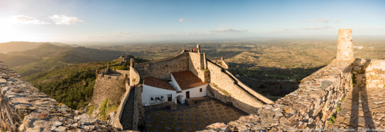 High view over thick stone ramparts into a small courtyard with a whitewashed red-roofed building and distant plain.