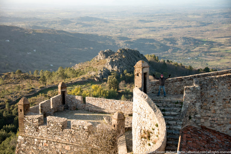 Stone castle walls, stairs, and small roofed turrets descending toward a broad valley, with one visitor on the rampart.