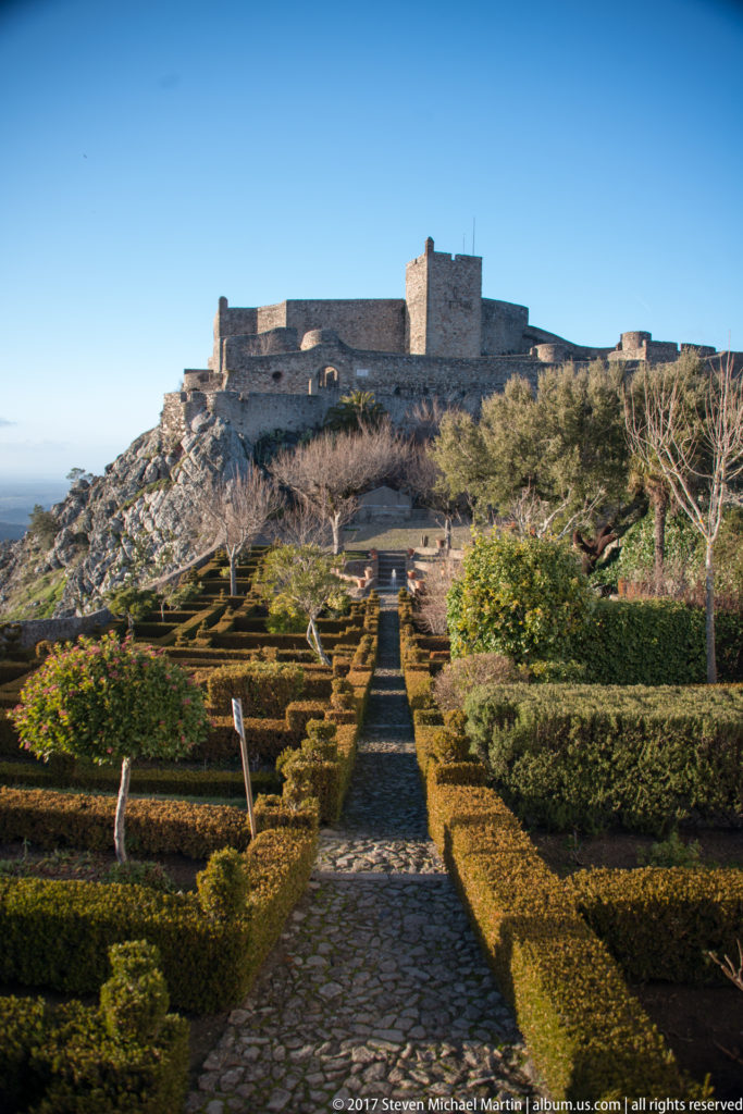 Cobbled garden path between clipped hedges leading toward the stone walls and square keep of Marvão Castle.