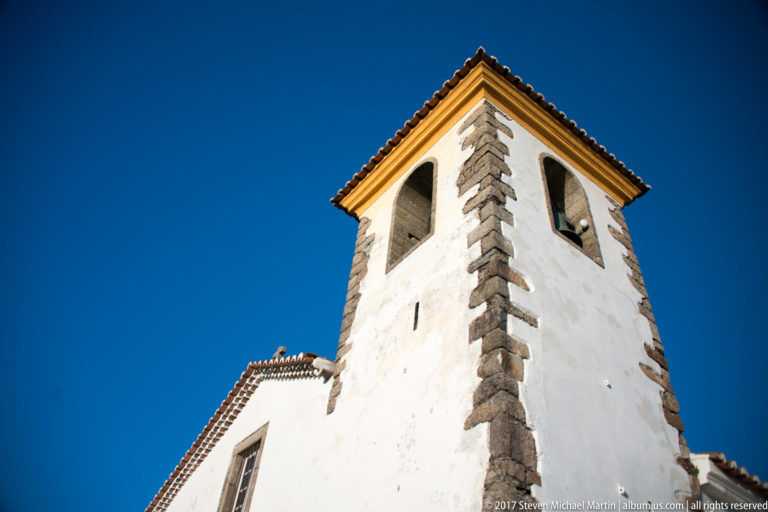 Low-angle view of a white church bell tower with gray stone corner blocks, arched openings, and tiled roof.