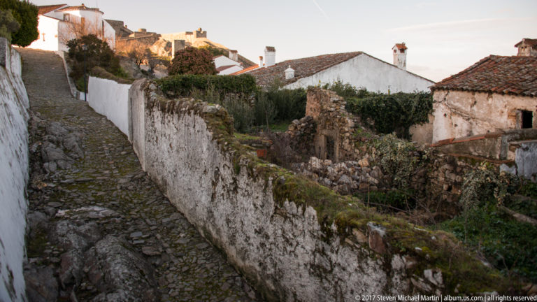 Narrow cobbled lane passes whitewashed walls and an overgrown garden beneath tiled roofs and chimneys.