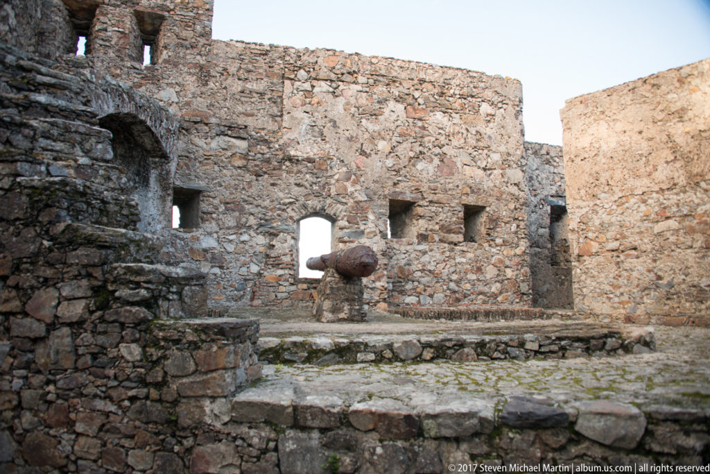 Weathered stone courtyard with wall openings, an arched recess, raised paving, and a dark spherical display object.