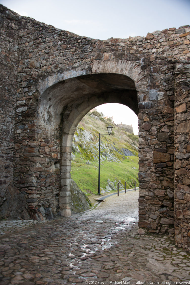 Wide stone arch opens from a cobbled fortified passage onto a grassy rocky slope with a lamp post.