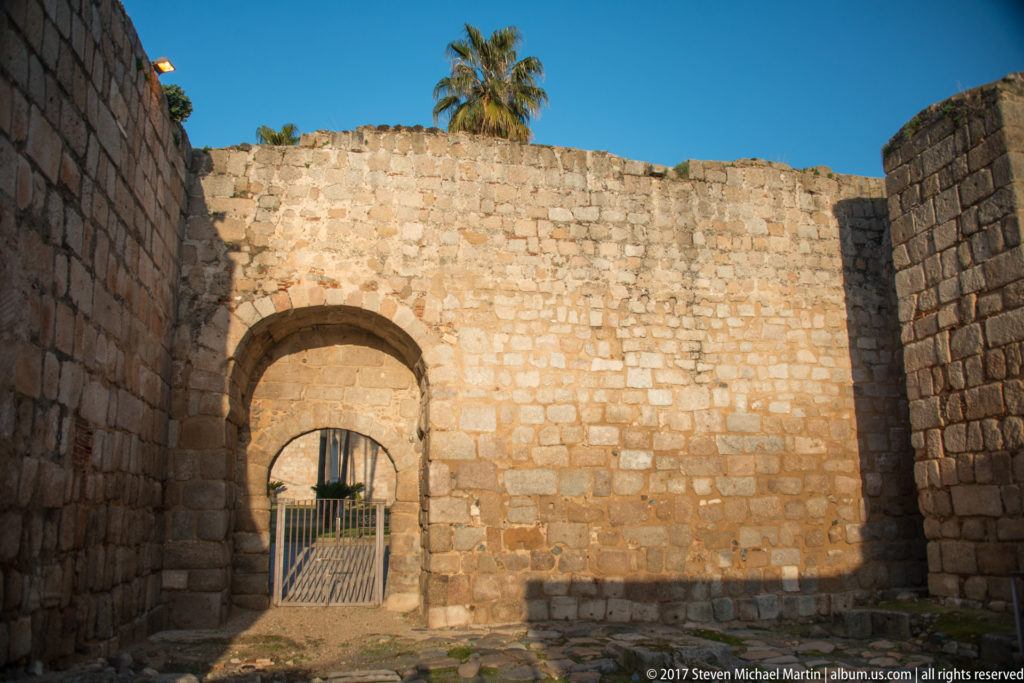 Stone arched gateway set into thick masonry walls, with a metal gate and palm tree visible beyond.