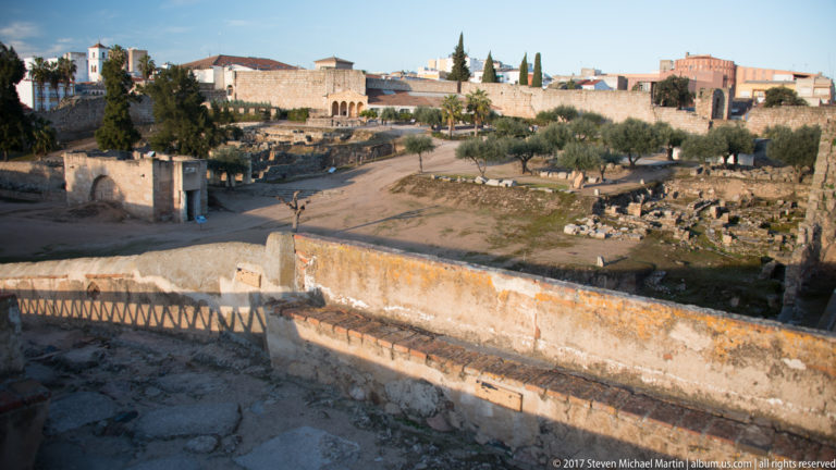 Elevated view over low ruins and olive trees enclosed by a high stone fortification wall.