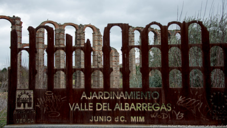 Rust-colored sign reading “Ajardinamiento Valle del Albarregas” in front of the arches of Mérida's Roman aqueduct.