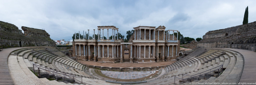 Wide elevated view of Mérida's Roman Theatre with curved stone seating and the monumental columned stage façade at center.