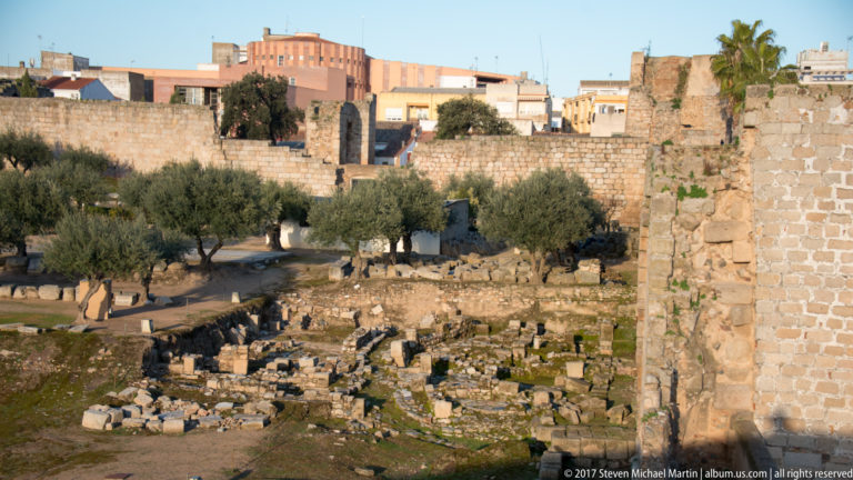 Low excavated stone foundations and olive trees beside a tall surviving fortification wall.