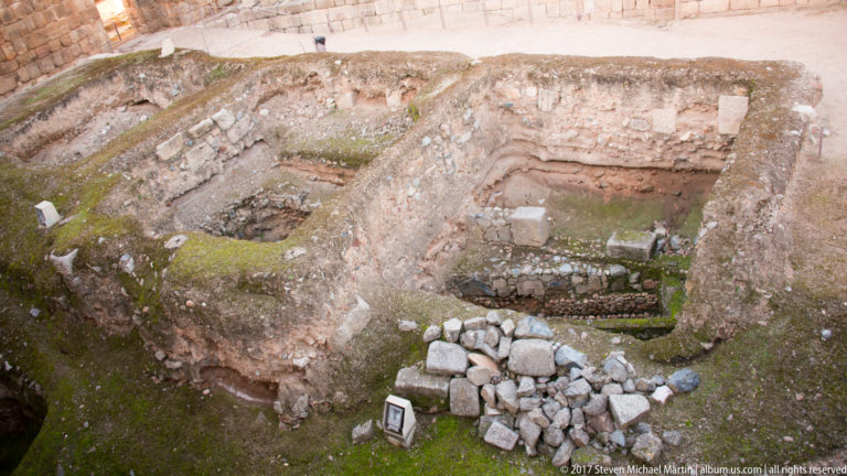Overhead view into a sunken excavation with thick walls, curved masonry, loose blocks, and moss.