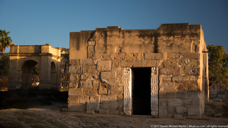 Small rectangular stone building with a dark doorway framed by carved panels in warm evening light.