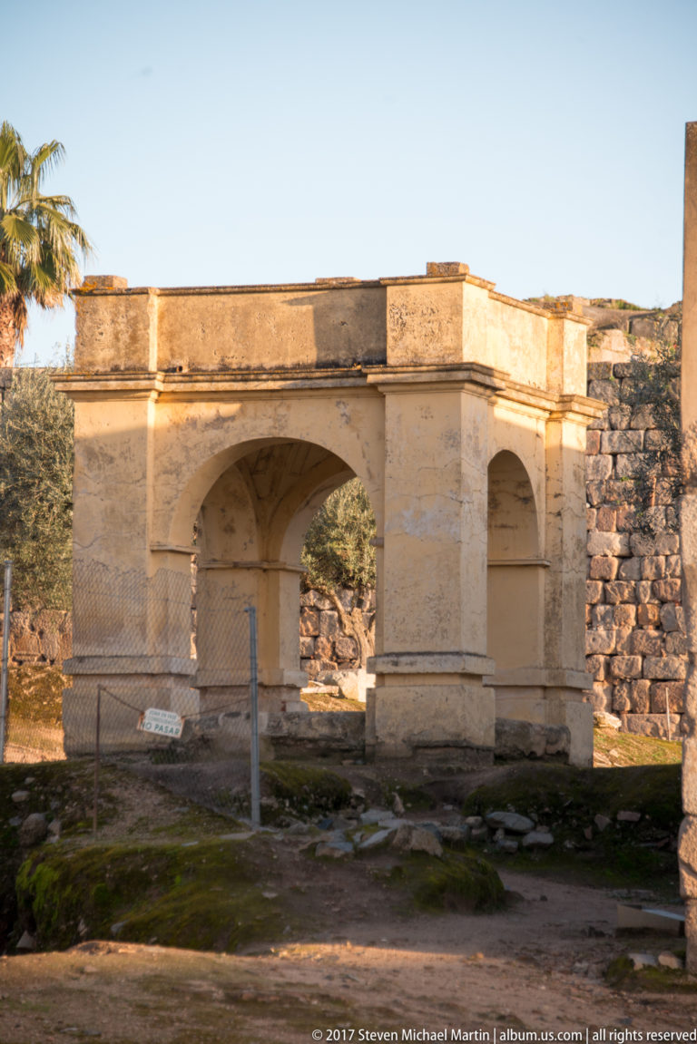 Pale two-arched masonry structure with heavy piers standing before a rough stone wall.