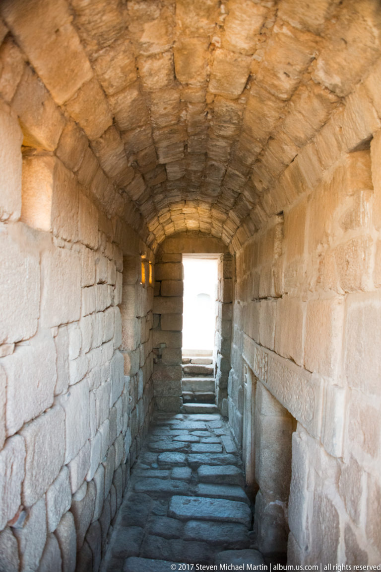 Long stone barrel-vaulted corridor with block walls and slab paving leading to a bright exit.