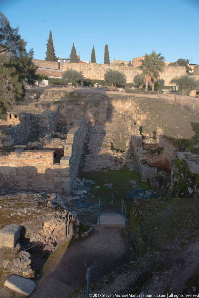 Broad archaeological area with parallel low walls and foundations below a high perimeter wall and trees.