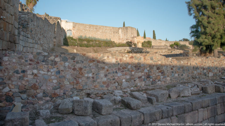 Excavated low stone walls and block foundations below a high fortified perimeter wall.