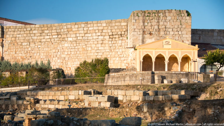 Pale three-arched portico with slender columns before a massive stone wall and excavated foundations.