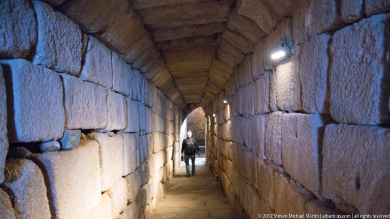Narrow stone-block corridor with low heavy ceiling, wall lights, and a visitor walking toward the exit.