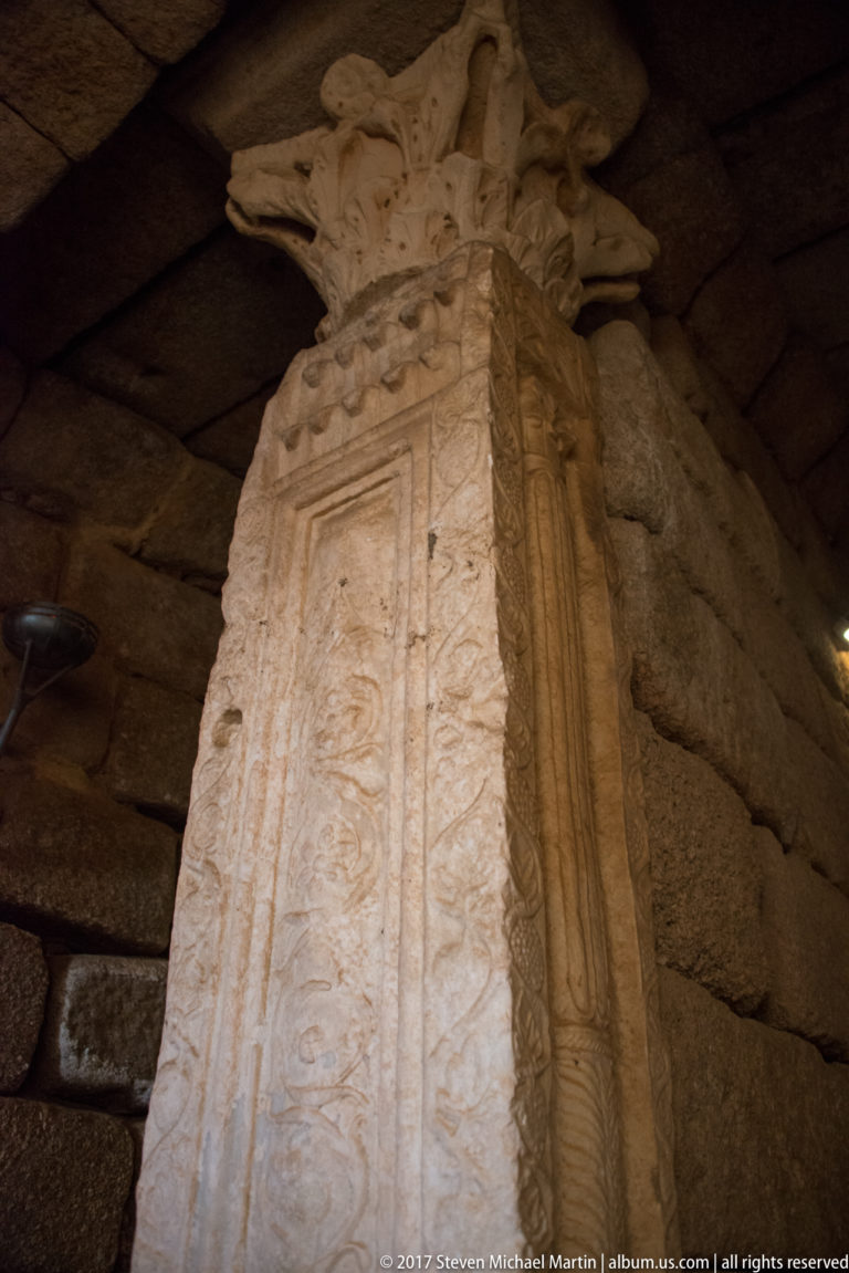 Tall carved stone support with relief ornament and projecting capital beneath a low stone ceiling.