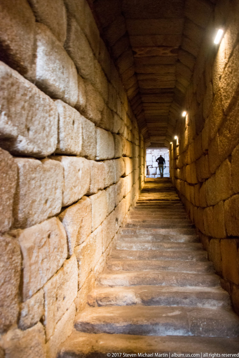 Long low stone corridor with slab floor and wall lights receding toward a bright opening.