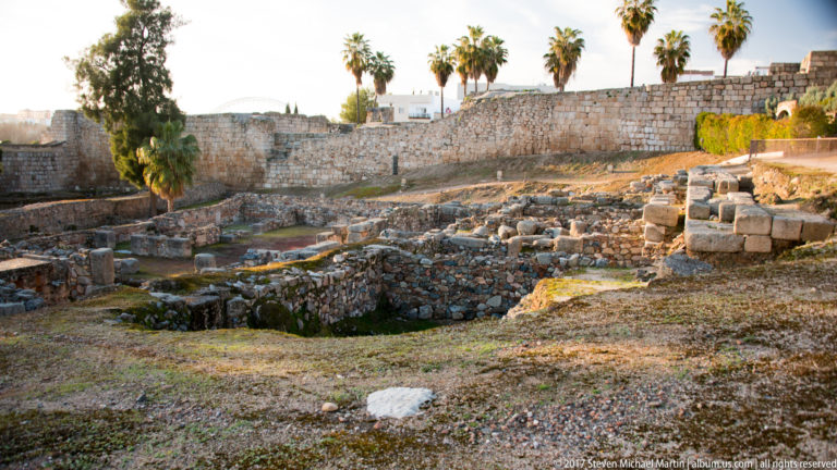 Low excavated stone foundations and trenches across open ground beneath a high wall and palm trees.