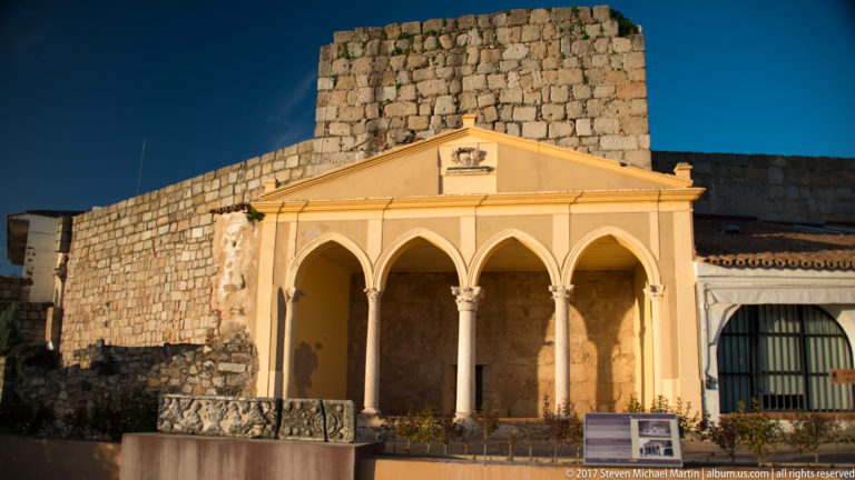 Pale gabled portico with three pointed arches and slender columns before a massive stone wall.