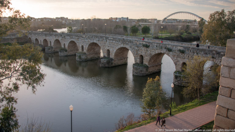 Long stone Roman bridge with multiple arches crossing the Guadiana, with pedestrians and a modern bridge behind.