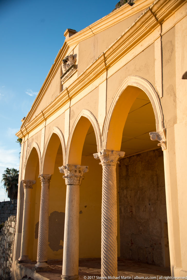 Oblique close view of pale arches on carved capitals and textured columns beneath a triangular pediment.