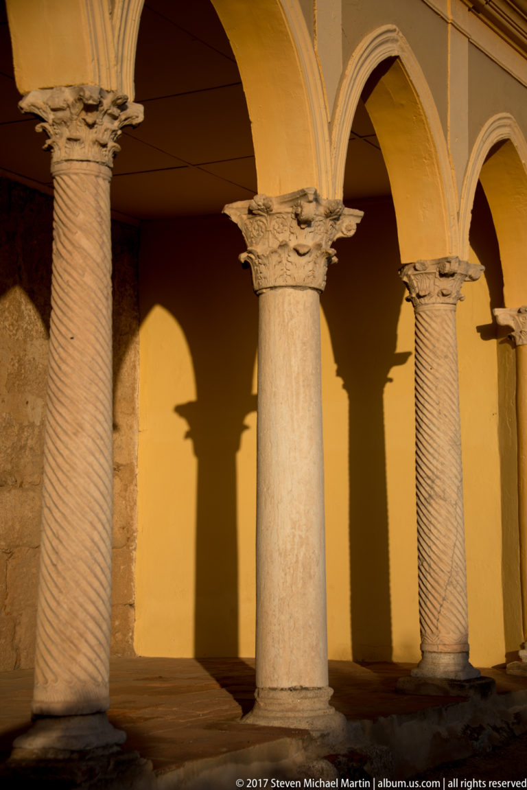 Spiral-fluted stone columns with carved capitals support rounded arches, casting long shadows across an ochre wall.