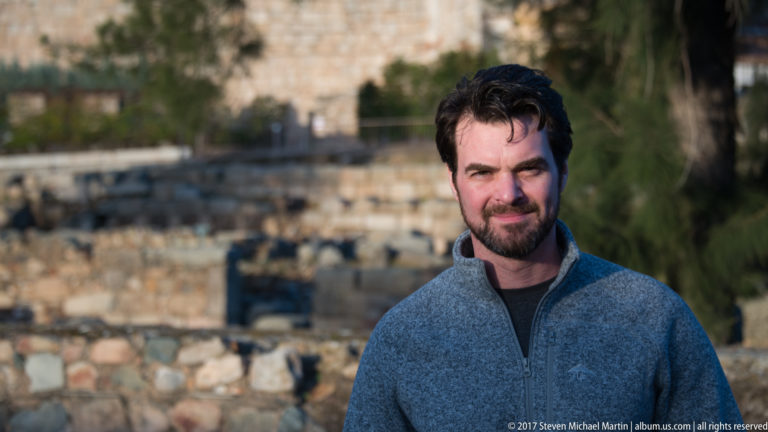 Bearded man in a gray sweater facing the camera, with archaeological stone remains blurred behind him.