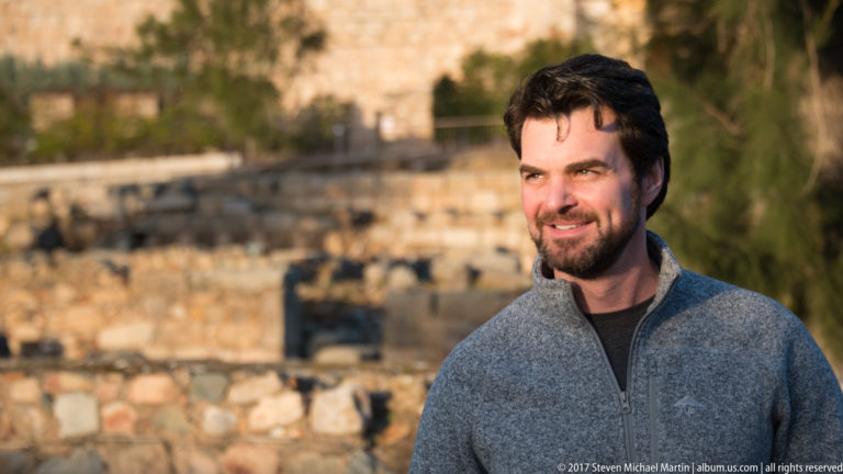 Smiling bearded man in three-quarter view, warmly lit against blurred stone archaeological remains.