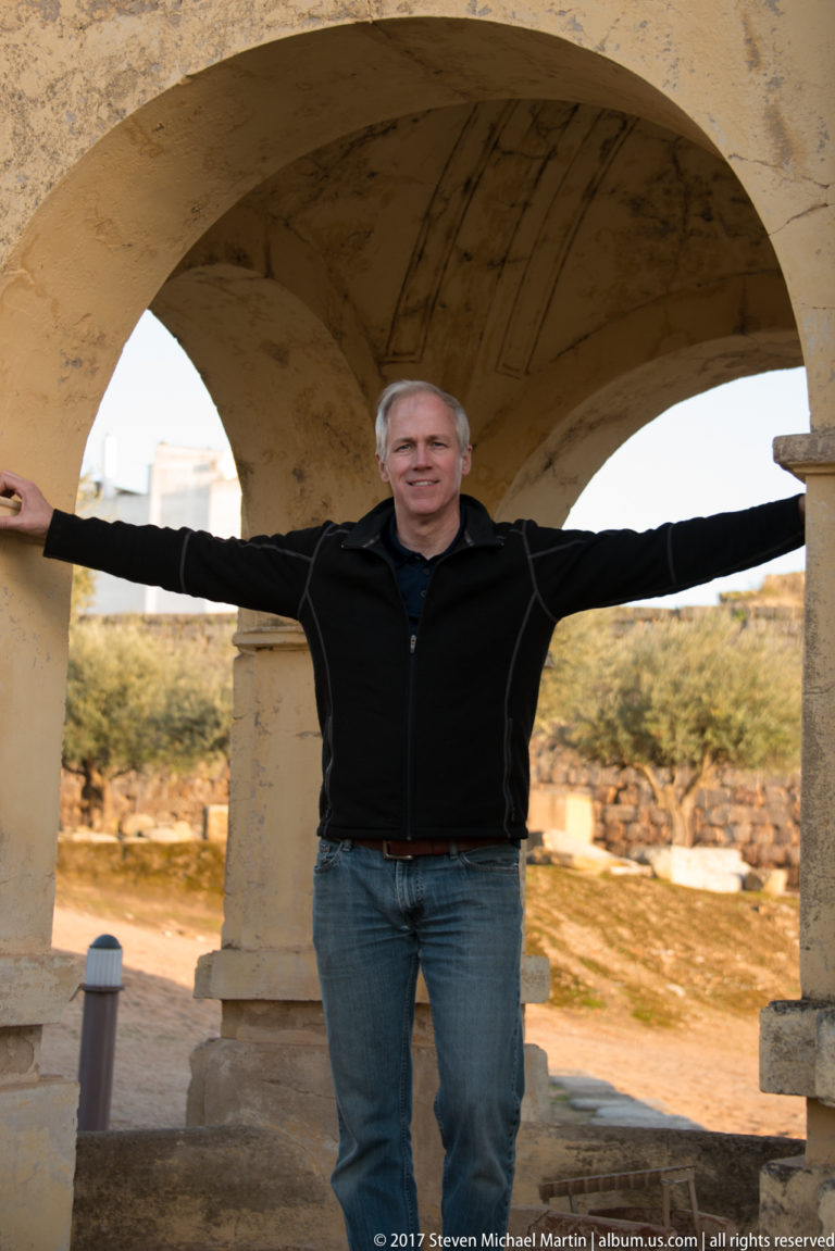 Man with arms outstretched beneath a broad stone arch, with archaeological remains visible beyond.