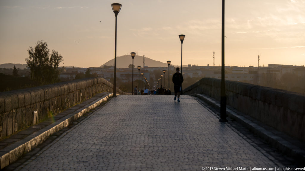 Long paved roadway of Mérida's Roman bridge at sunset, lined with stone parapets, lampposts, and pedestrians.