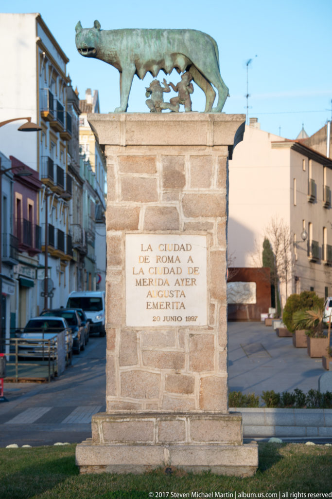 Stone sister-city monument topped by the Capitoline Wolf, with an inscription linking Mérida and Rome.