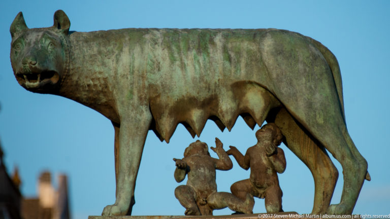 Bronze Capitoline Wolf in profile with the infant twins Romulus and Remus beneath her against blue sky.