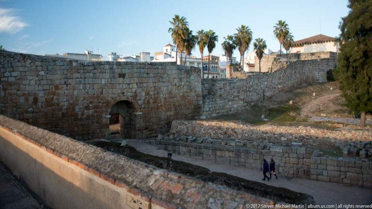 High stone fortification wall with a small arched opening, archaeological remains below, and palms above.