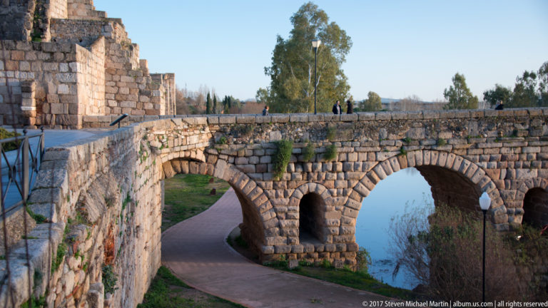 Stone arches of Mérida's Roman bridge above a curving riverside path, with pedestrians visible on the parapet.