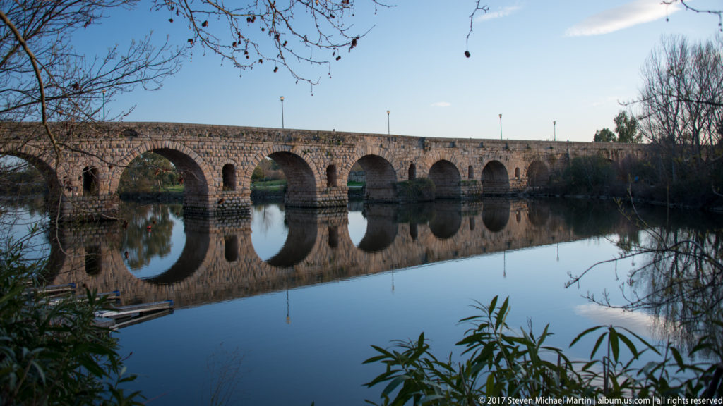 Long side view of Mérida's Roman bridge, its repeated arches mirrored almost perfectly in still river water.