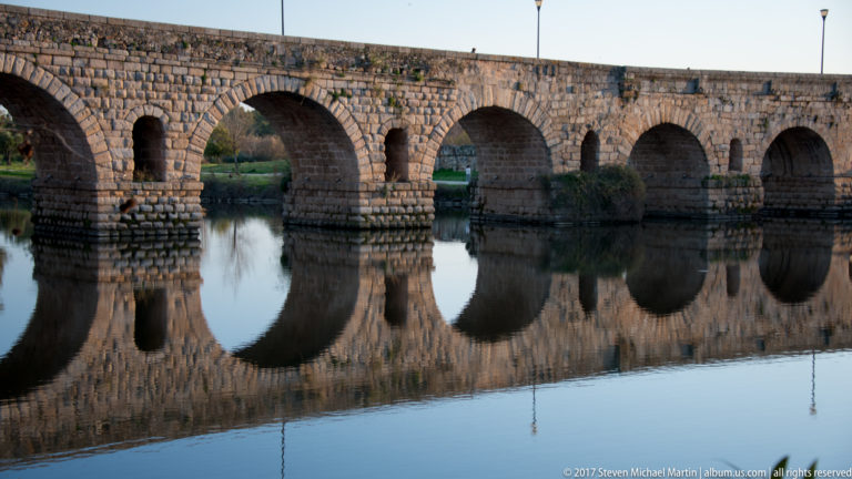 Several stone arches and heavy piers of Mérida's Roman bridge reflected in smooth river water.