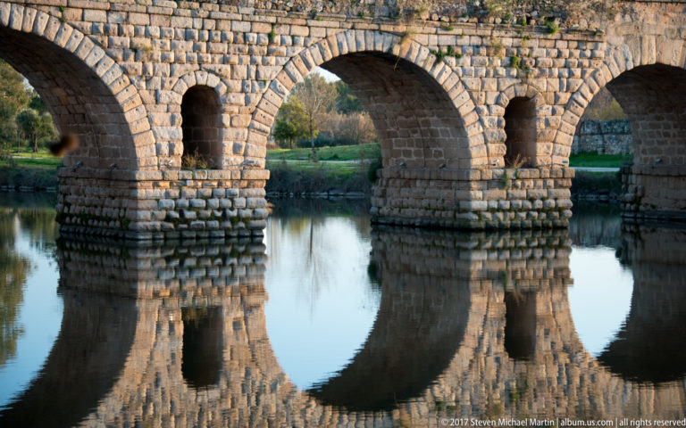 Close view of three stone bridge arches and piers reflected in the Guadiana, forming near-circular shapes.