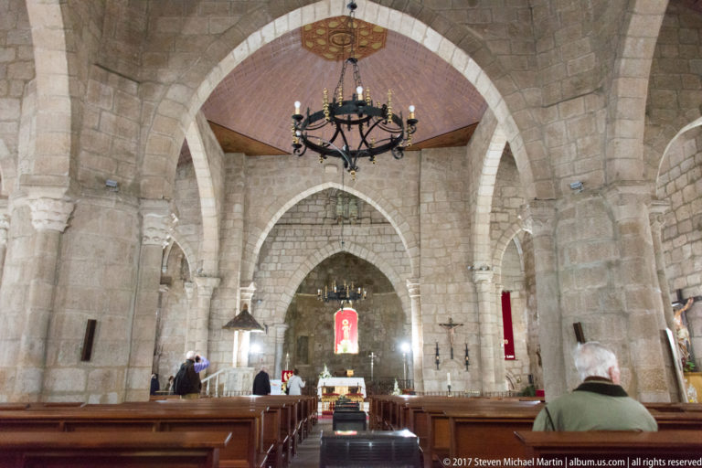 Stone nave of the Basilica of Santa Eulalia with broad arches, wooden pews, chandelier, and sanctuary beyond.