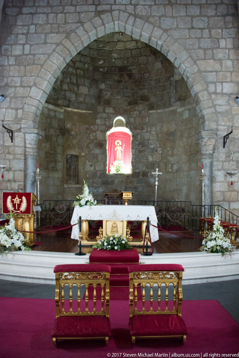 Frontal view of a church sanctuary with white altar, flowers, candles, red textiles, and broad stone arch.