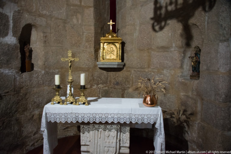 Small side altar with crucifix, candles, lace cloth, gilded tabernacle, devotional figures, and rough stone wall.