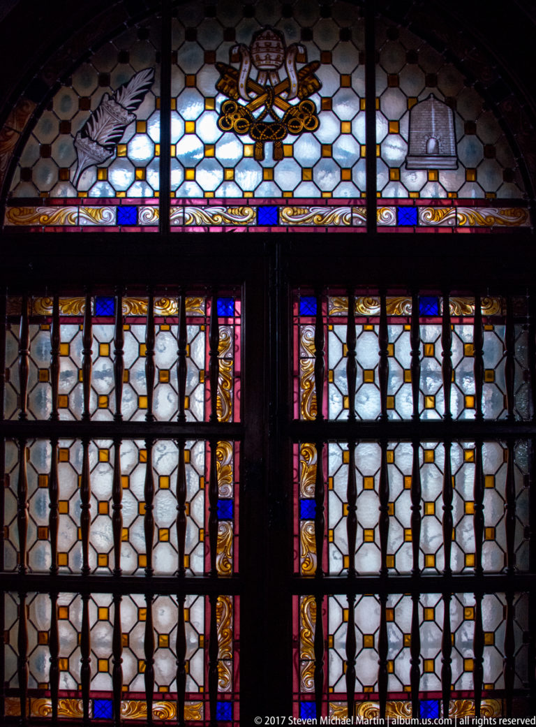Colorful stained-glass church window seen through a dark metal protective grille with geometric latticework.