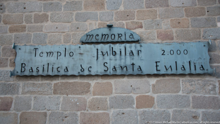 Stone plaque reading “Memoria Templo Jubilar 2000 Basílica de Santa Eulalia” mounted on a block masonry wall.