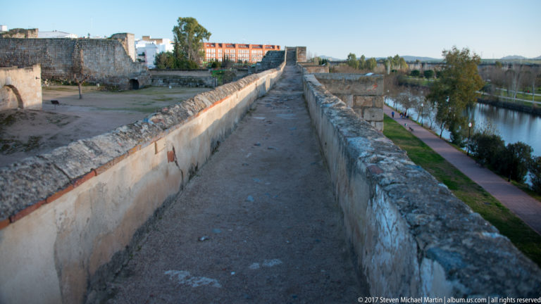 Narrow elevated passage between stone parapets, with the river and riverside path visible at right.