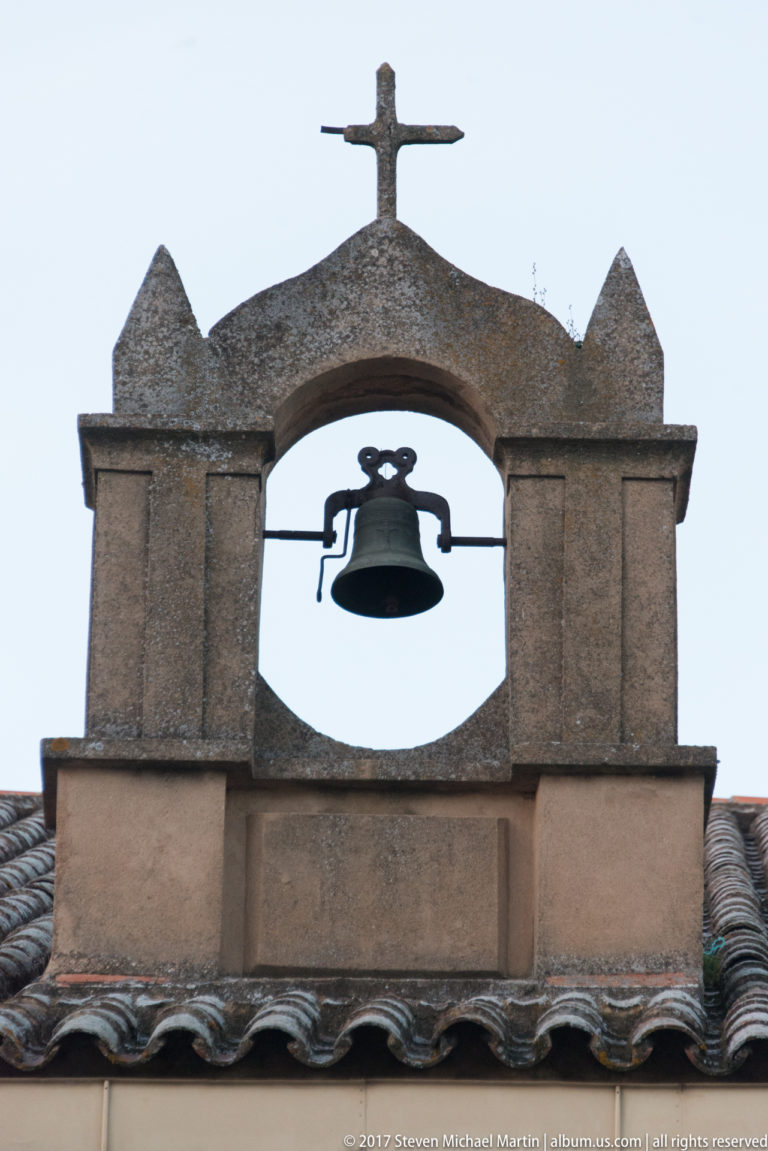 Single bell hanging in an arched masonry bell gable topped by a cross above a tiled roof.