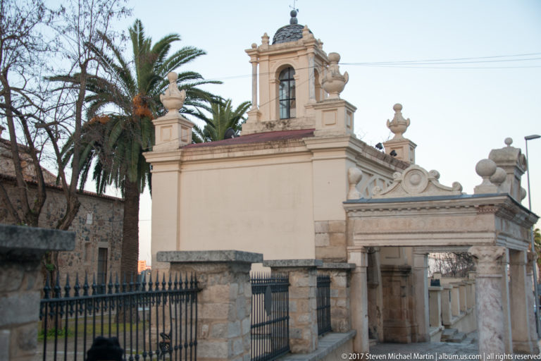 Ornate pale entrance with small bell opening, finials, scrollwork, iron gates, and palm trees beside older stone walls.