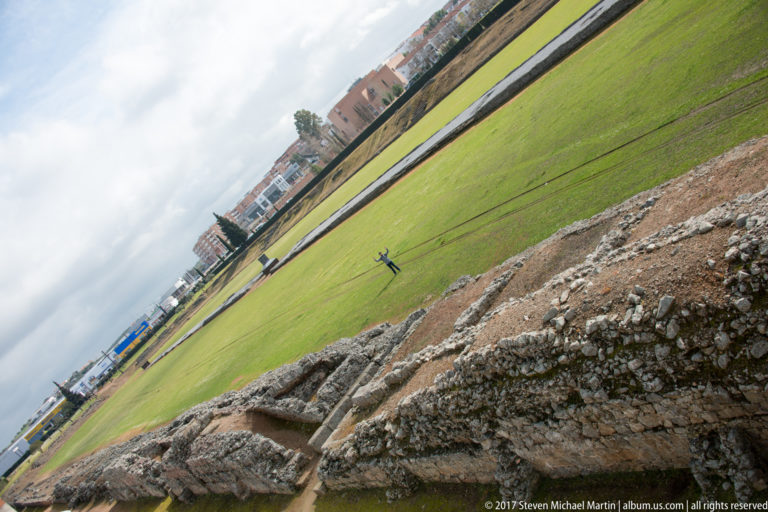 Wide grassy arena of Mérida's Roman circus seen past rough masonry, with long central remains and city buildings beyond.