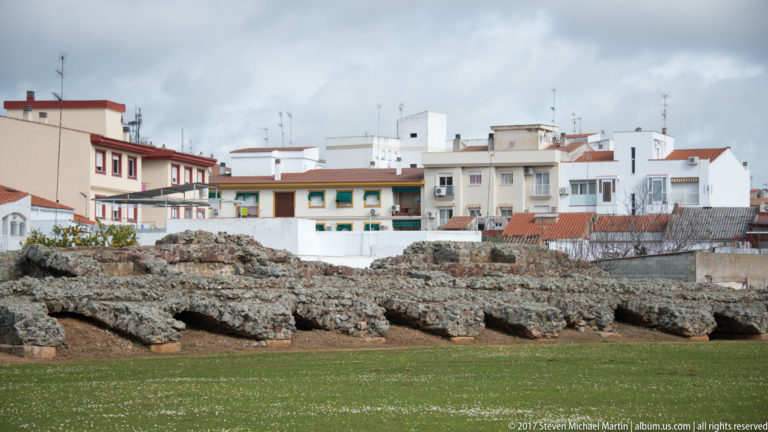 Low weathered masonry remains beside the grassy Roman circus site, with modern residential buildings immediately behind.