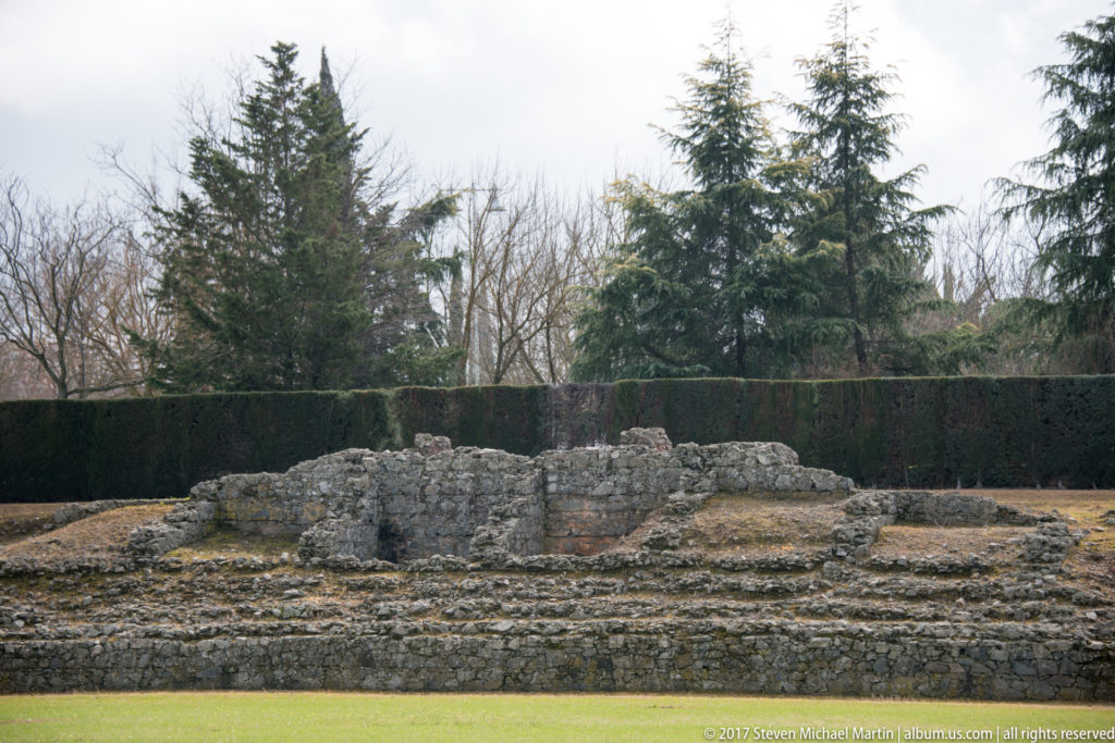 Low excavated stone foundations forming several compartments beside the Roman circus grounds, backed by hedges and trees.